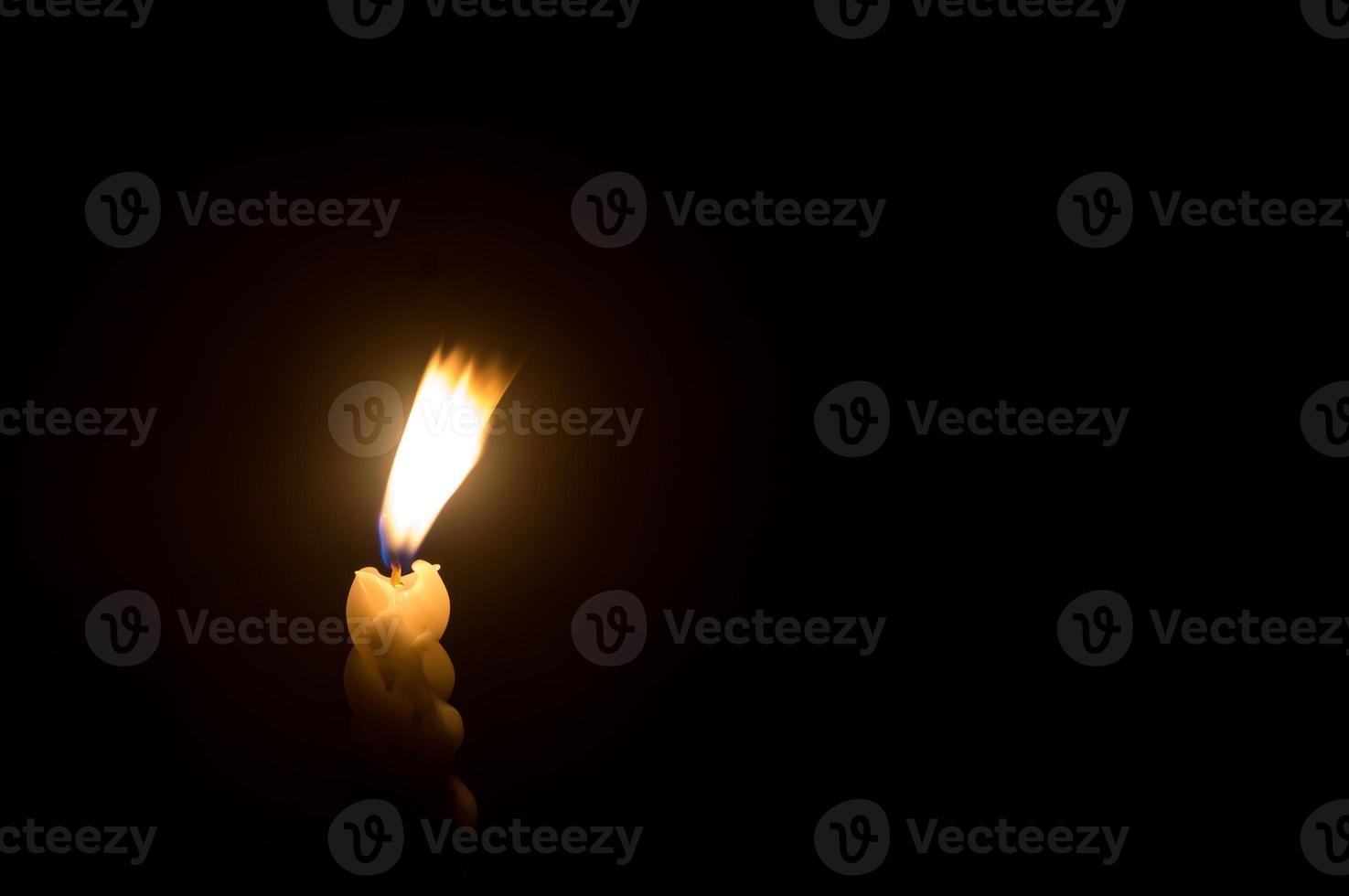 Blurred photo of single burning candle flame or light glowing on a spiral white candle against blowing wind on black or dark background on table in church for Christmas, funeral or memorial service