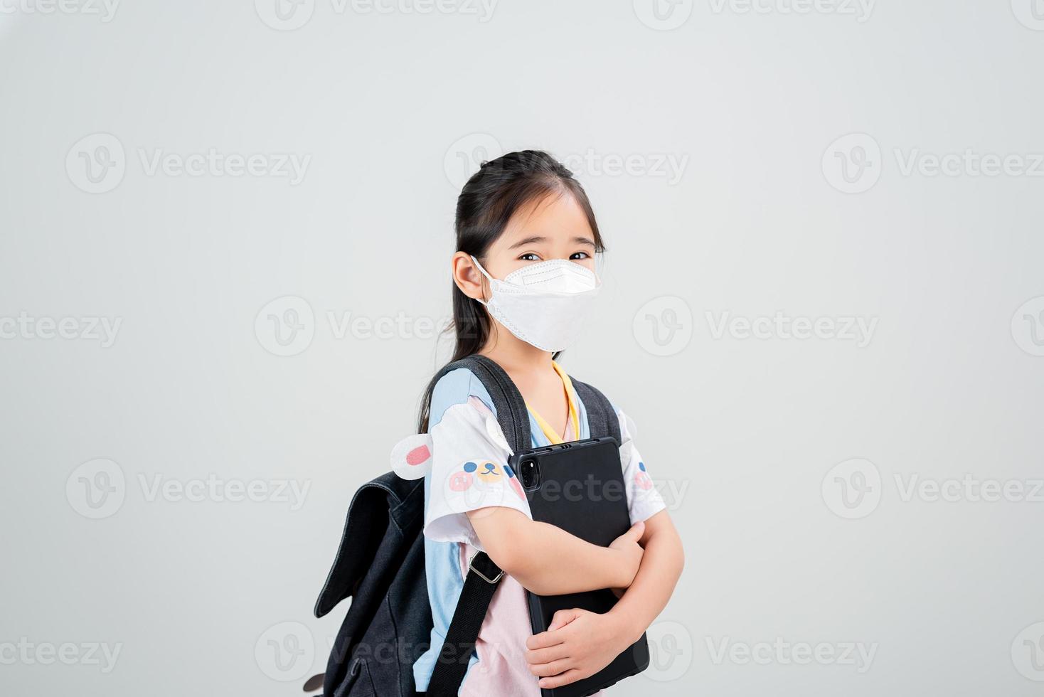 Asian little child girl carry a backpack wearing respirator mask to protect coronavirus outbreak and pointing hand to blank background, New virus Covid-19 photo