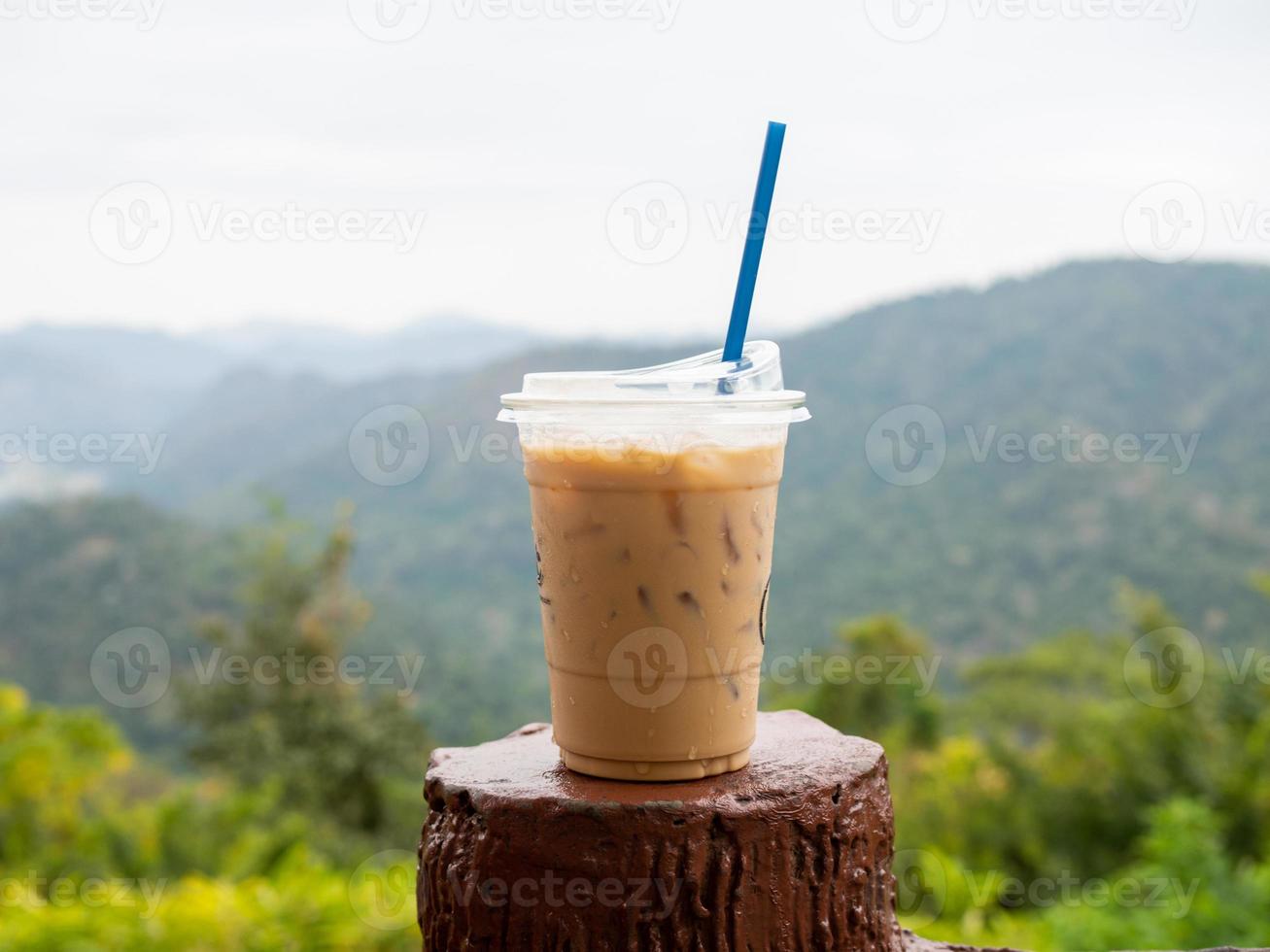A glass of iced coffee is placed on the fence against a backdrop of mountains and sky. photo