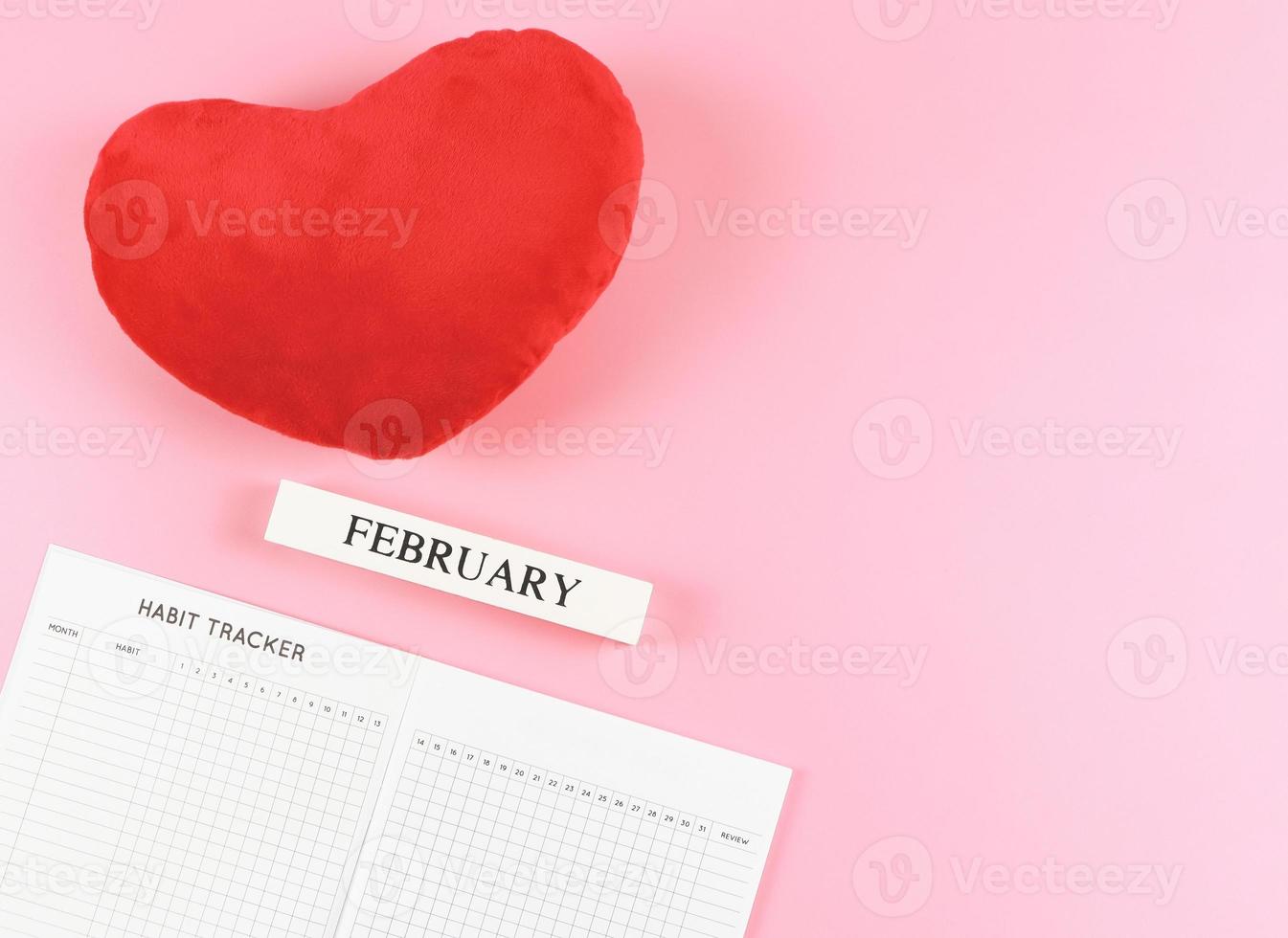 lat lay of habit tracker book, wooden calendar February,  red heart shape pillow on pink  background with copy space. photo