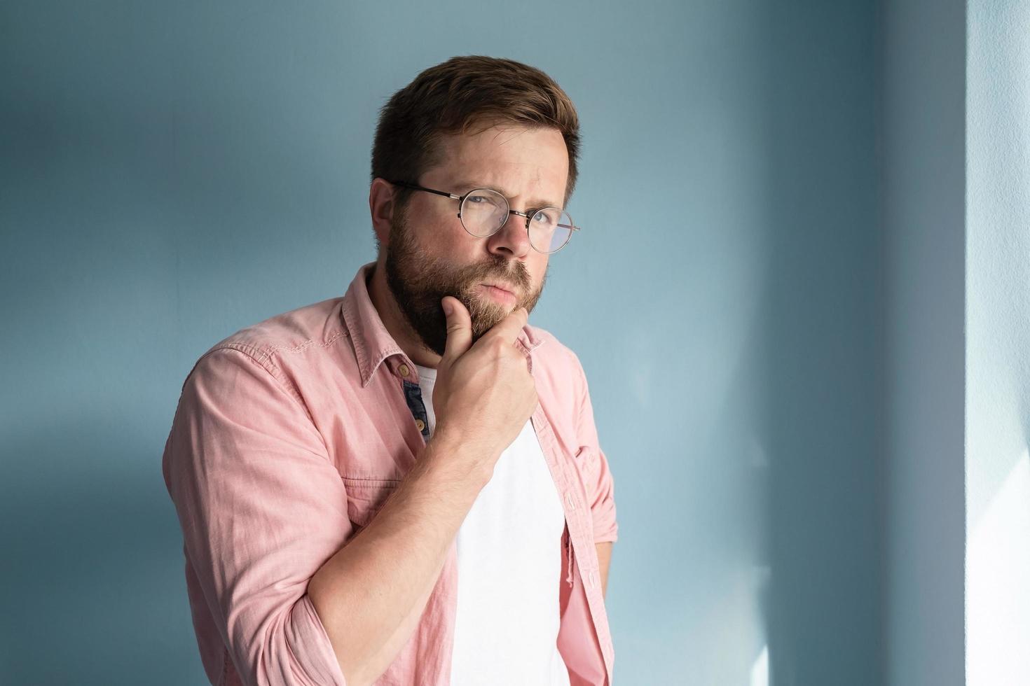 Caucasian man in glasses looks incredulously, suspiciously with a squint, thinking about something and touches his beard with hand. photo