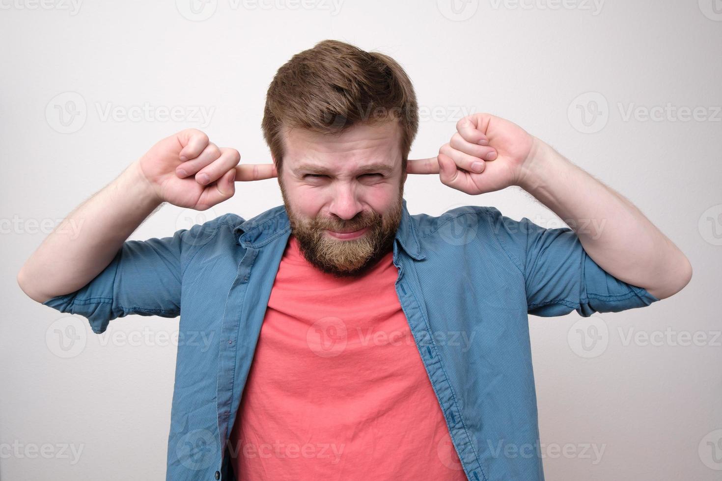 Displeased, irritated Caucasian man tired of loud noise, covering ears with fingers and frowning at the camera. White background. photo