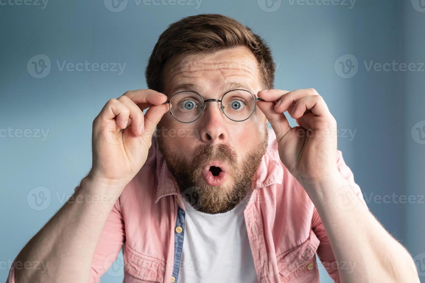 Man is shocked, he does not believe his eyes, he holds glasses with hands and opens mouth in surprise. Blue background. photo