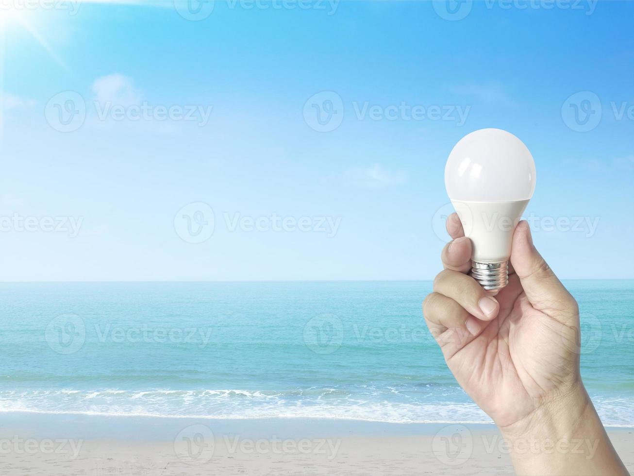 Hand holding a LED light bulb on a sandy beach with clear sky. Using economical and environmentally friendly light bulb concept photo
