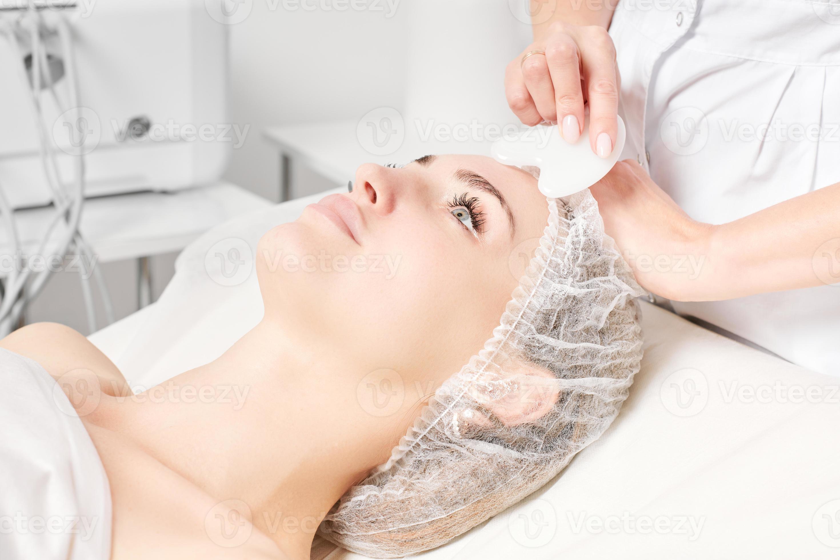 Cosmetologist making facial massage with Gua Sha stone of woman face