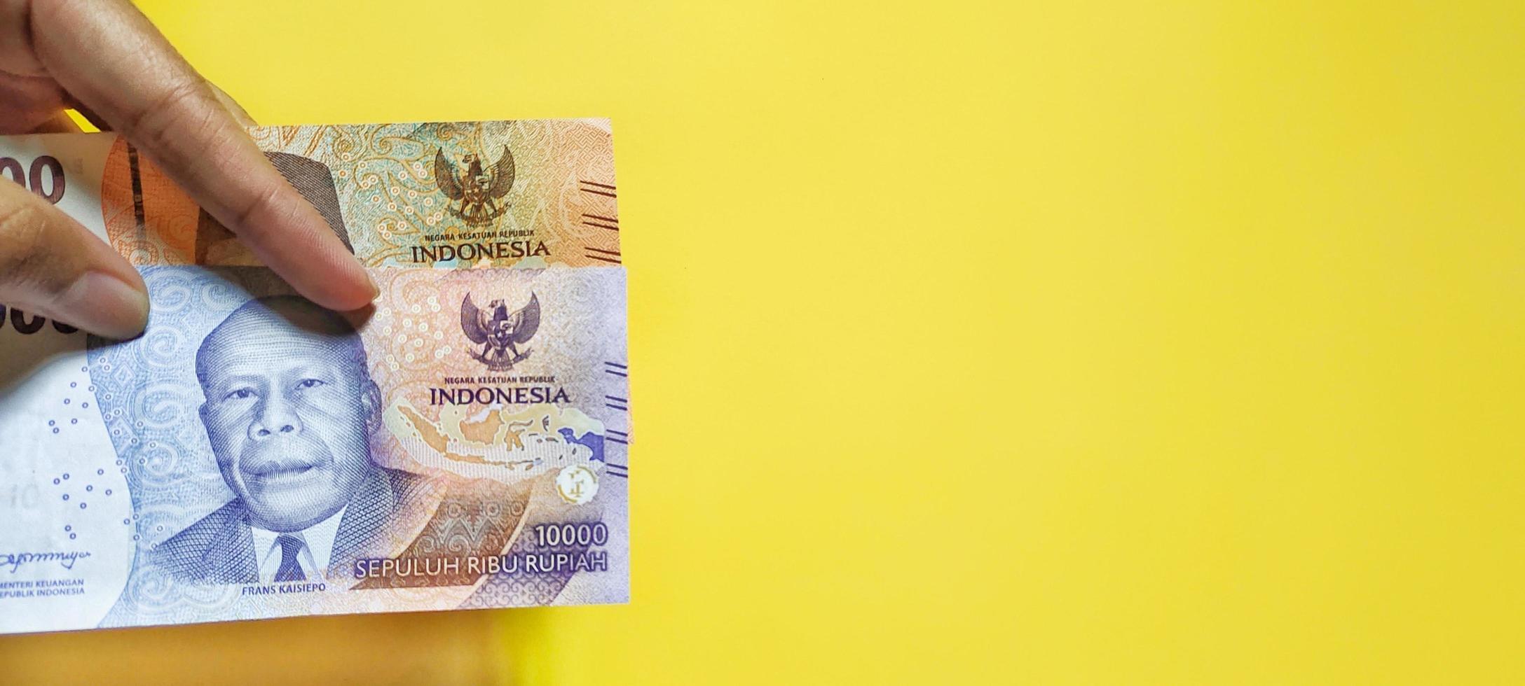 hombre que sostiene billetes en rupias indonesias de denominación 5000 y 10000 última edición, aislado en fondo amarillo. foto