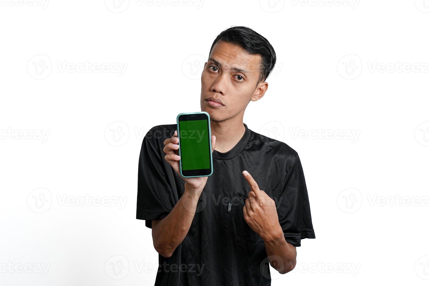 emocionado hombre asiático con camiseta negra de entrenamiento, apuntando a la pantalla verde del teléfono inteligente. aislado por fondo blanco foto