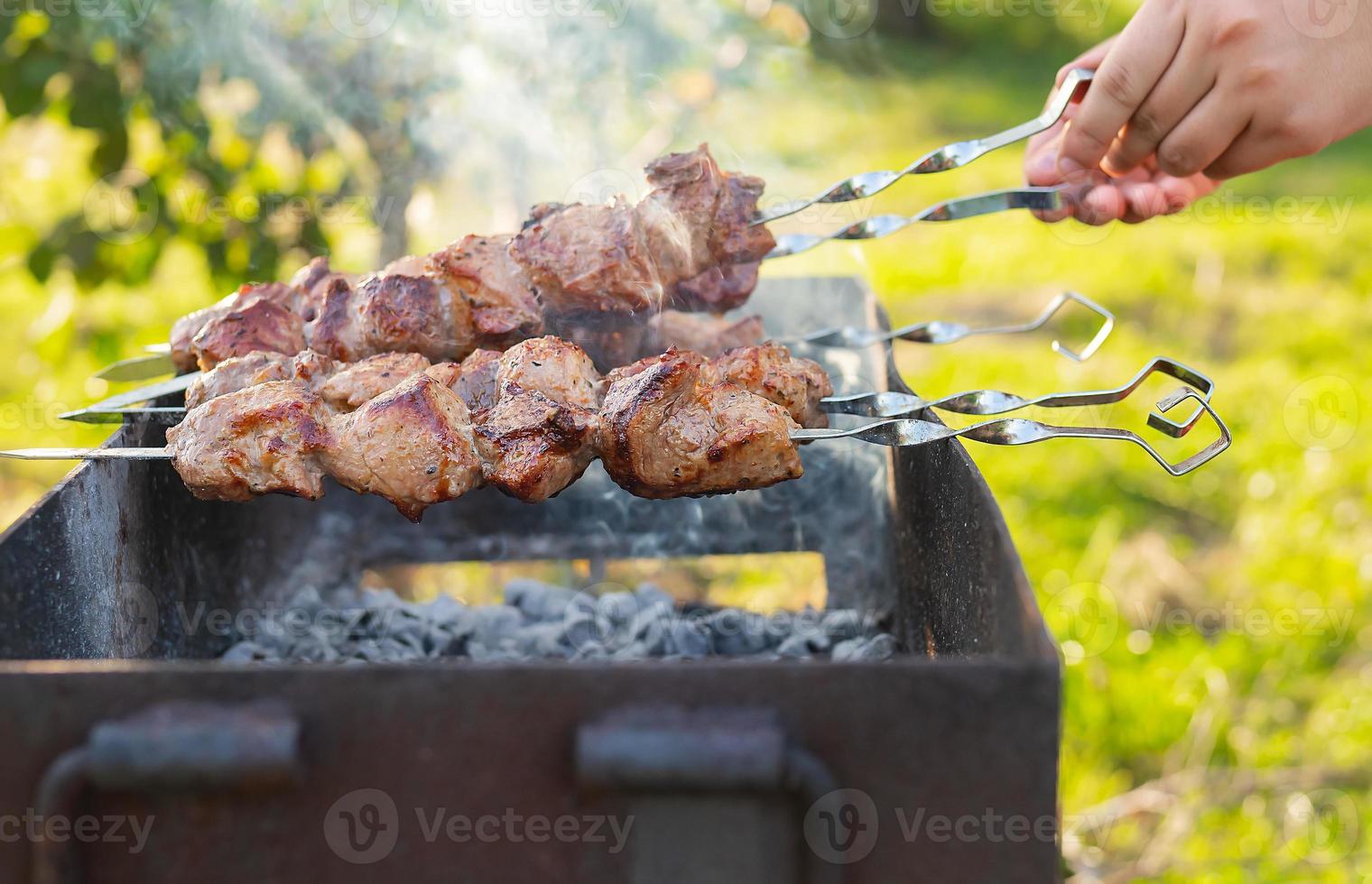 Cooking shish kebab on the grill with smoke, the man flips the skewers. Fresh meat for barbecue grilled on fire. Picnic in nature. photo