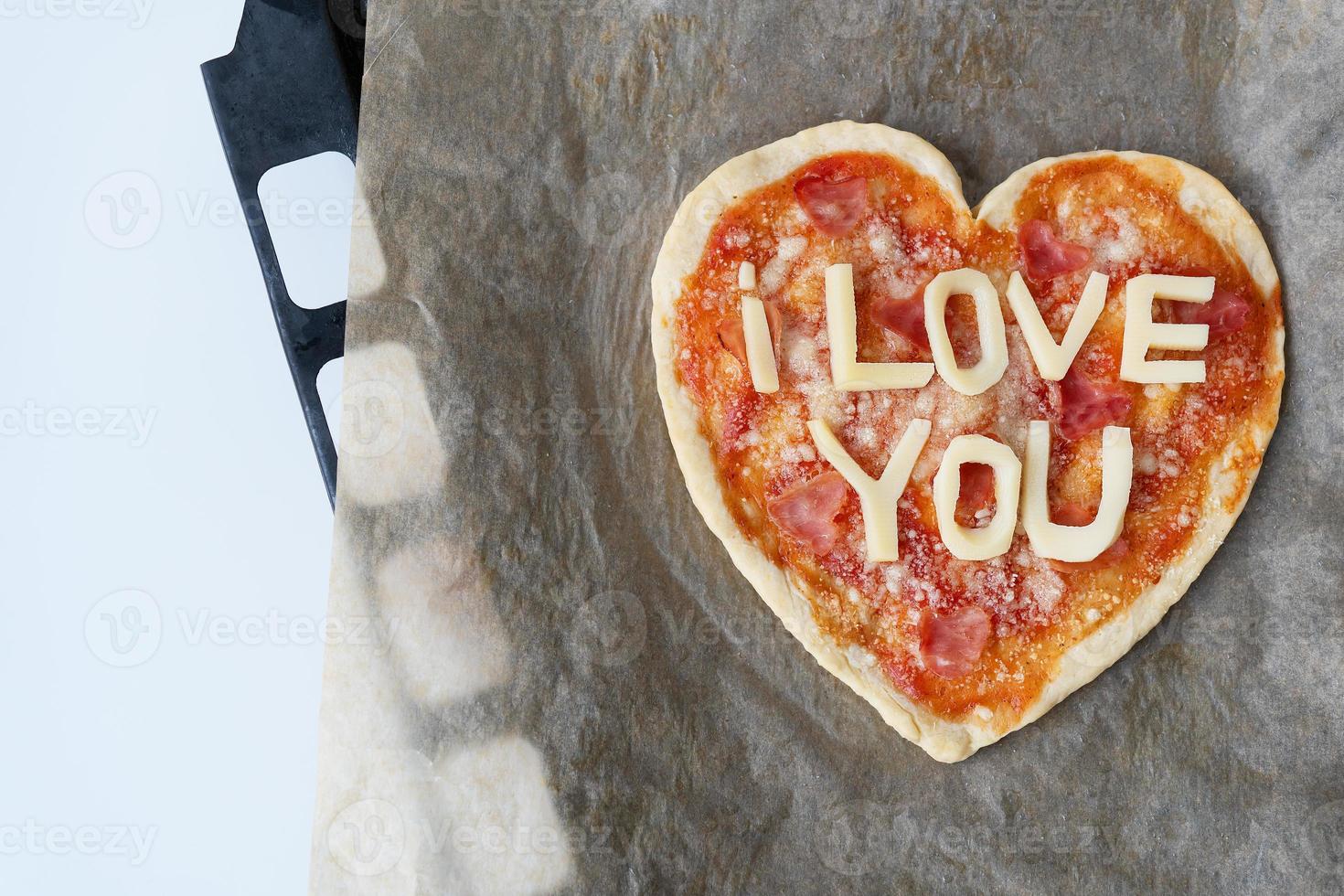 Italian heart shaped pizza with salami, tomato sauce, parmesan, pizza sauce, mozzarella and olive oil on parchment paper with cheese I love you. Love concept for Valentine's Day February 14. photo