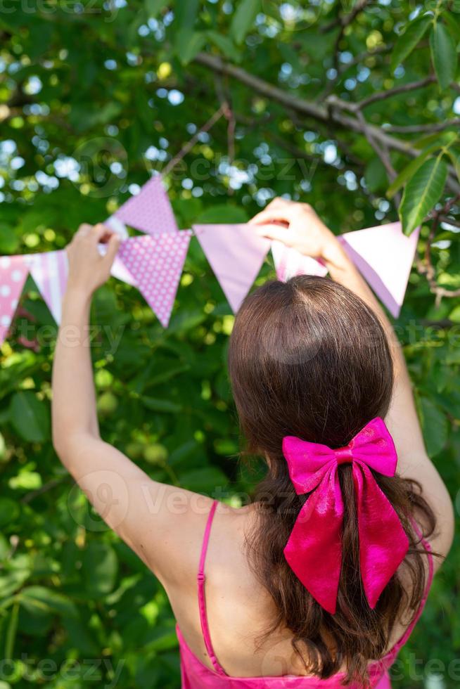 The girl hangs triangular flags of pink color against the backdrop of a green garden. A girl in a pink dress and a bow on her hair. Festive decor - celebration concept. photo