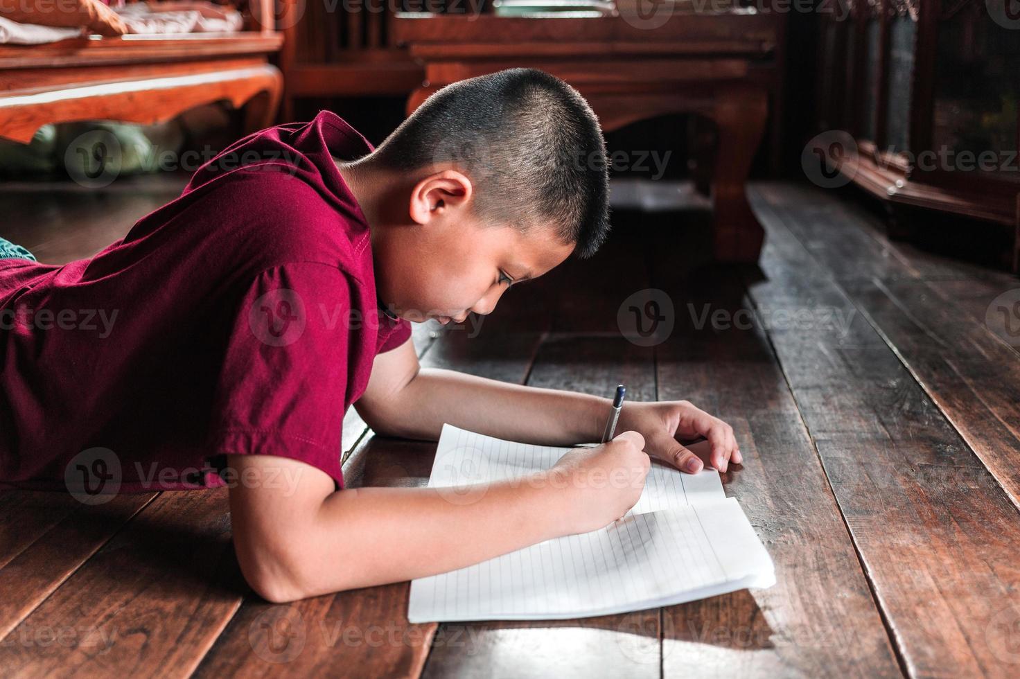 primer plano de un niño asiático sentado en el suelo de madera escribiendo un libro o haciendo deberes con una cara muy determinada en casa. foto