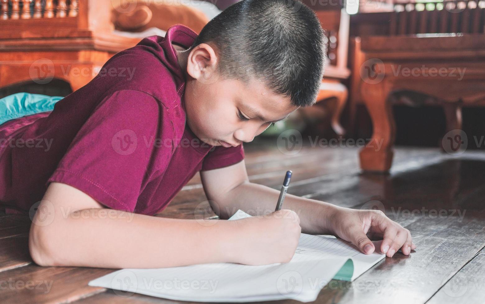 primer plano de un niño asiático sentado en el suelo de madera escribiendo un libro o haciendo deberes con una cara muy determinada en casa. foto