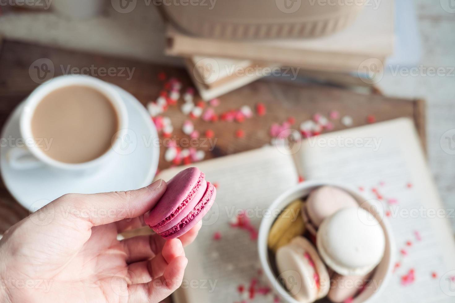 A hand holds a purple macaroon over a cup of coffee and a book. atmospheric photo