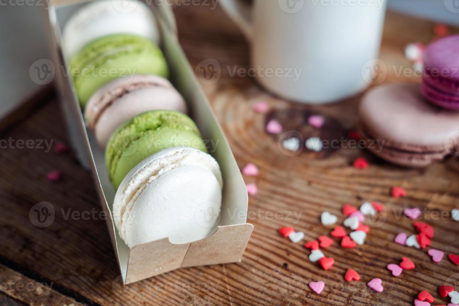 macarons en una caja, una taza de café, una jarra de leche contra el fondo de pequeños corazones sobre un fondo de madera, vista superior foto