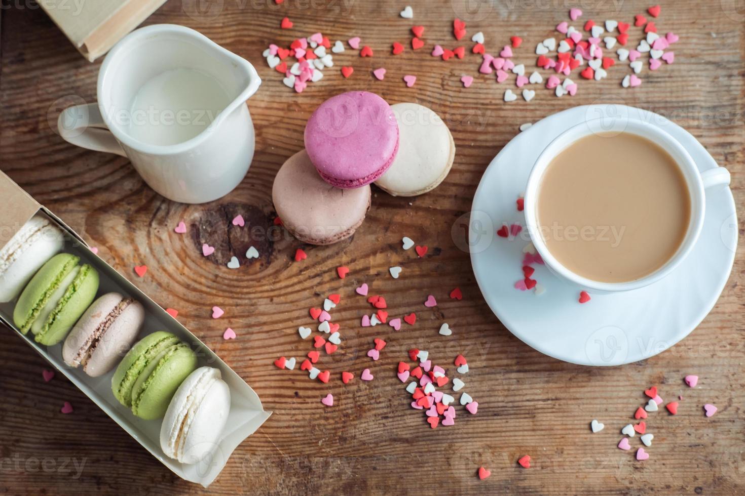 macarons en una caja, una taza de café, una jarra de leche contra el fondo de pequeños corazones sobre un fondo de madera, vista superior foto