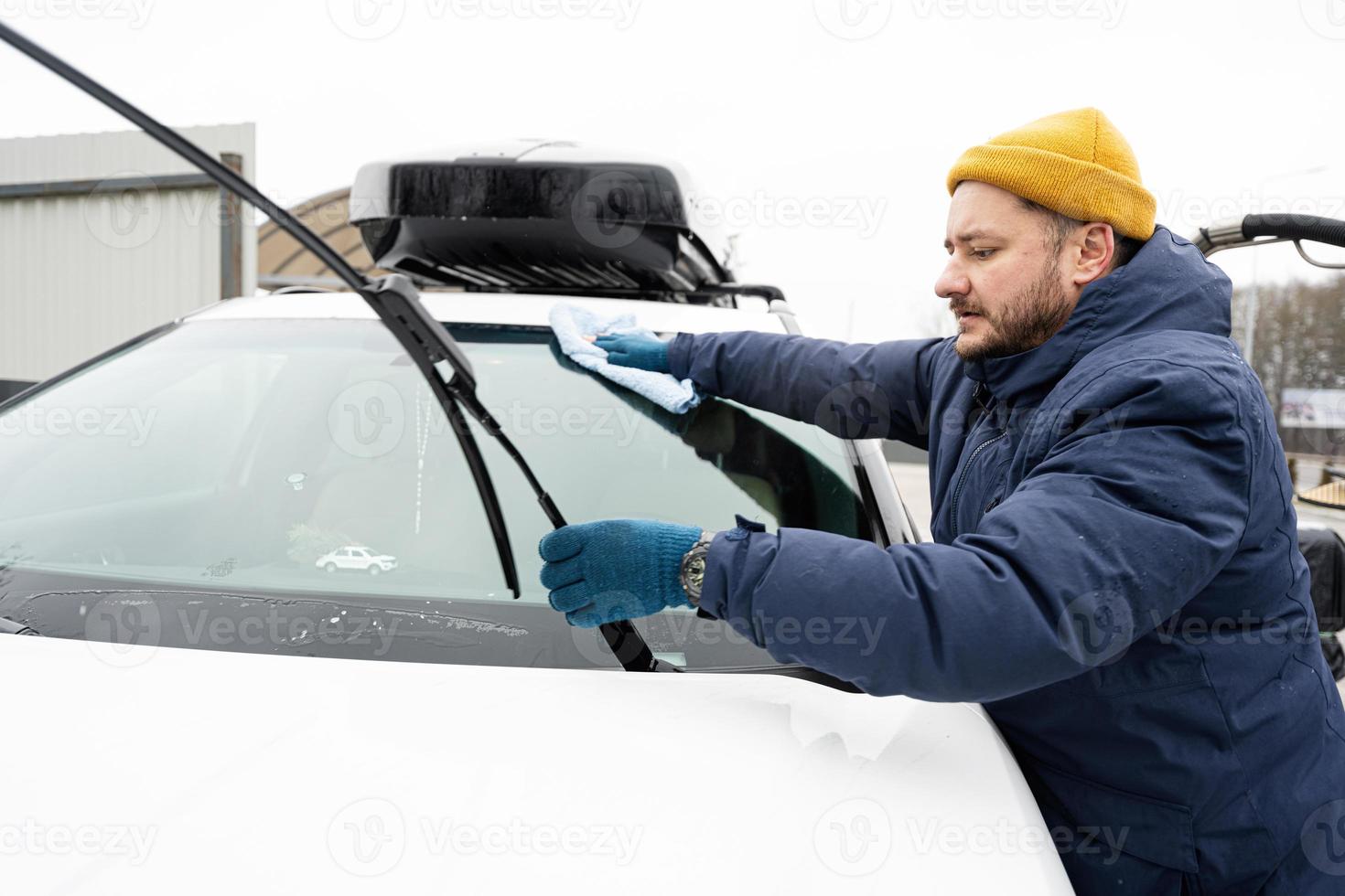 el hombre limpia el parabrisas de un vehículo suv americano con un paño de microfibra después de lavarlo en clima frío. foto