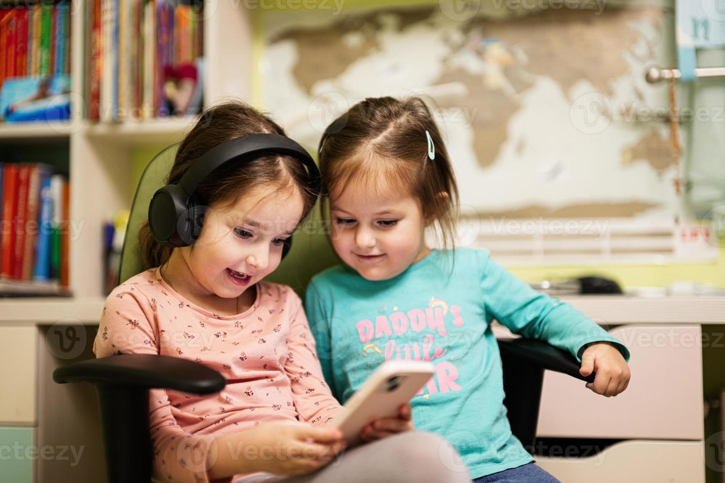 Two little girls sisters wear headphones watching cartoons or kid video on mobile phone. photo