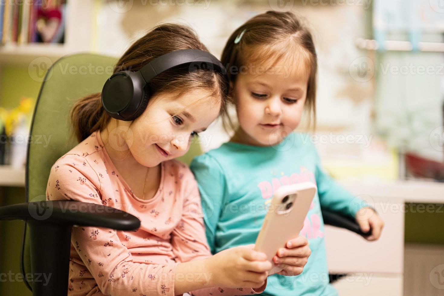 Two little girls sisters wear headphones watching cartoons or kid video on mobile phone. photo