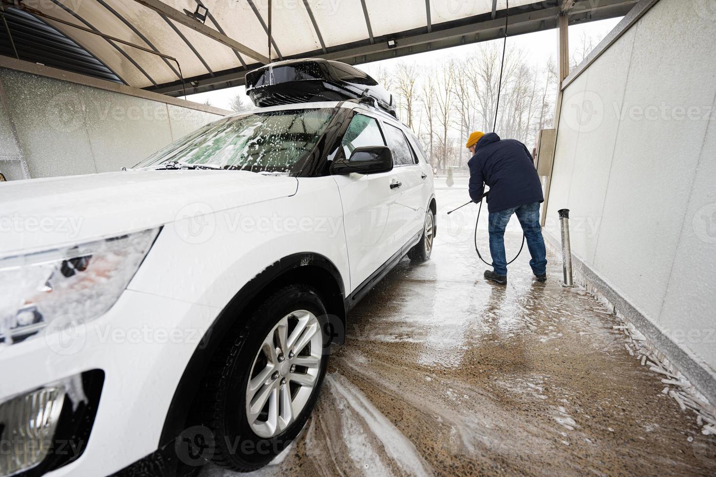 hombre lavando agua a alta presión coche suv americano con portaequipajes en autoservicio de lavado en clima frío. foto