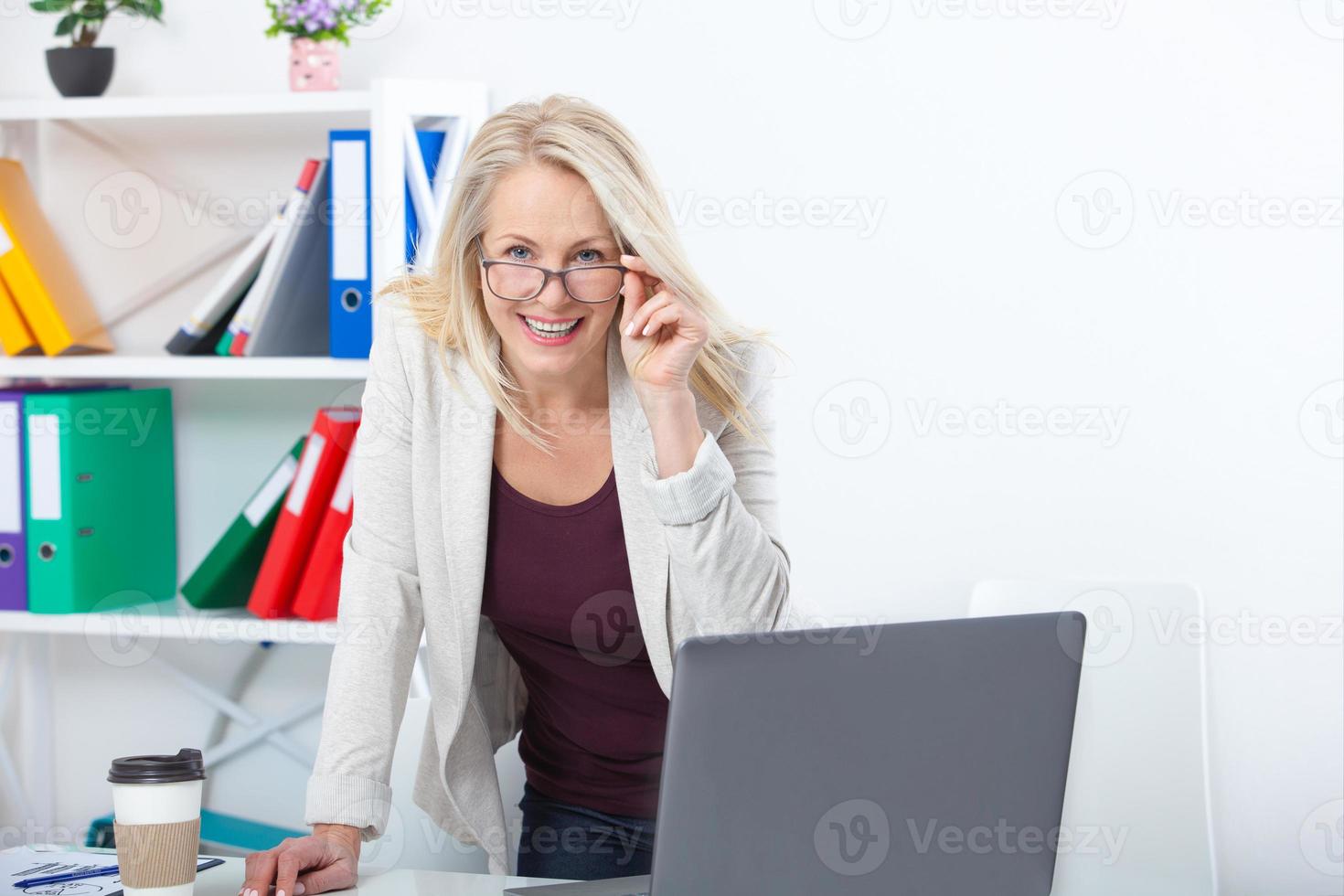 su trabajo es su vida. mujer de negocios con gafas trabajando en la oficina con documentos. hermosa mujer de mediana edad mirando a la cámara con una sonrisa mientras se sienta en la oficina. foto