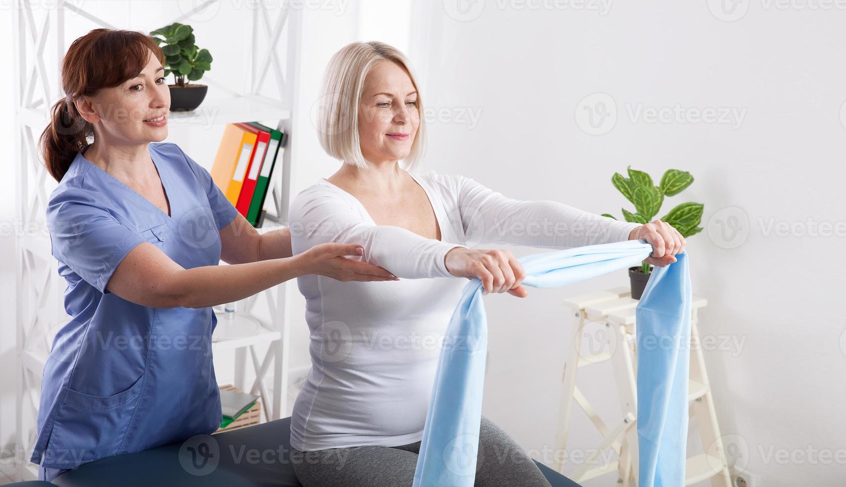 Physiotherapist and woman sitting on a bed exercising with a rubber tape photo