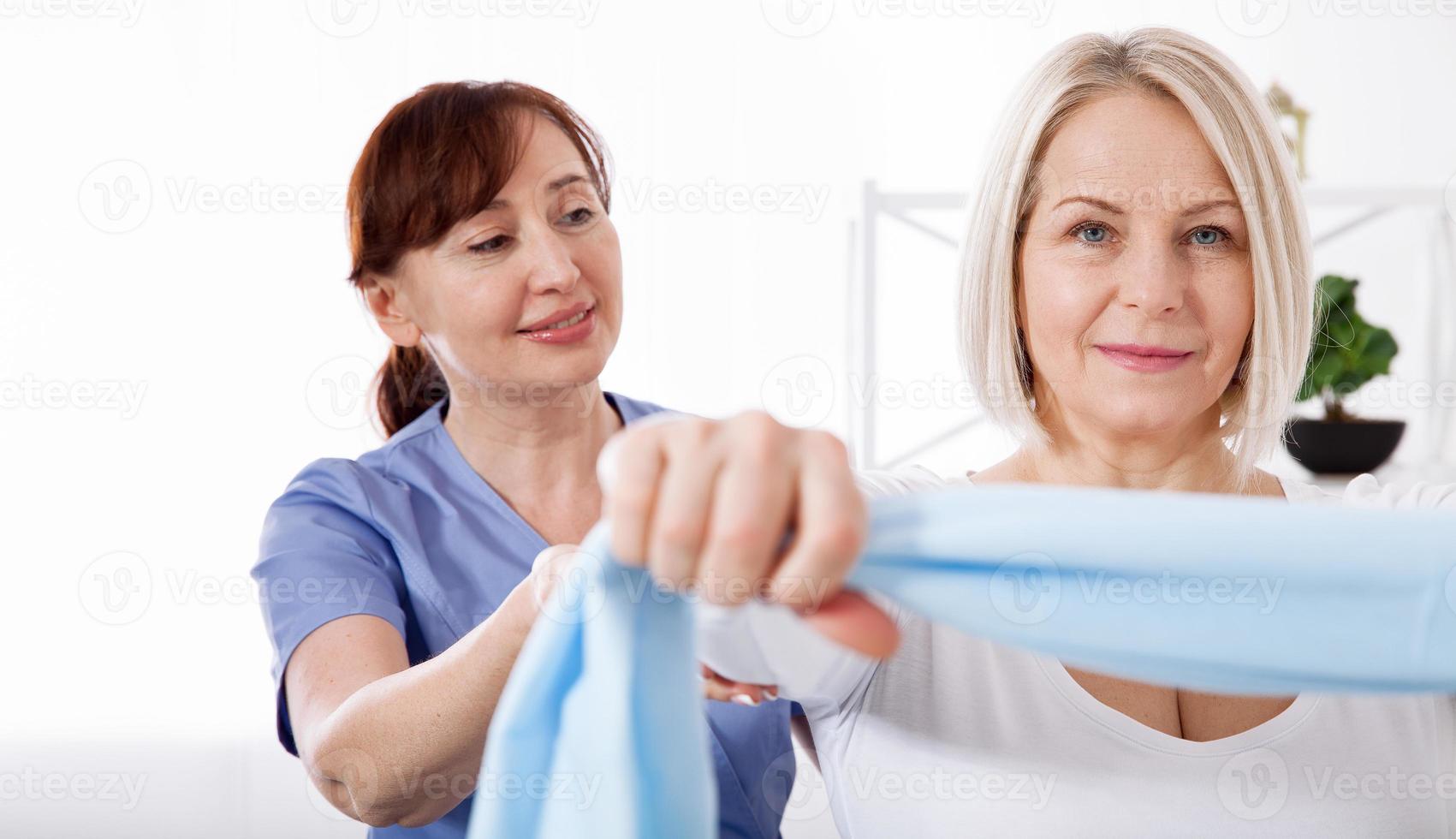 Physiotherapist and woman sitting on a bed exercising with a rubber tape photo