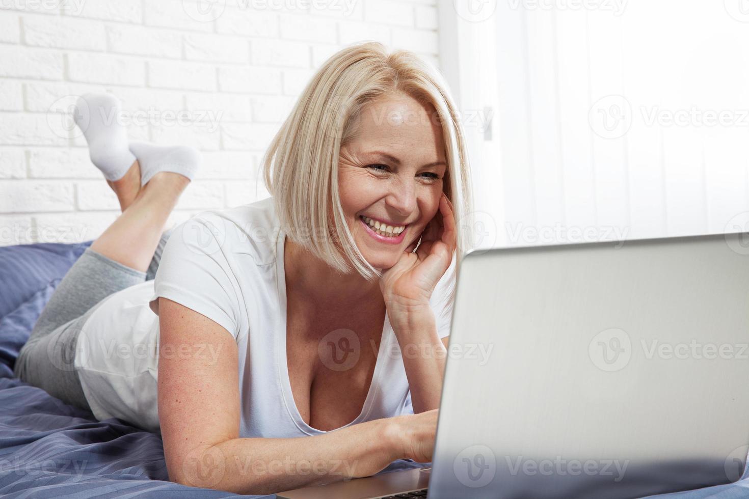 Pretty smiling woman has pleasant smile, haircut, dressed in casual domestic clothes, laugh during watching comedy, leaves comments, chats online with friends on laptop computer sits in bed photo