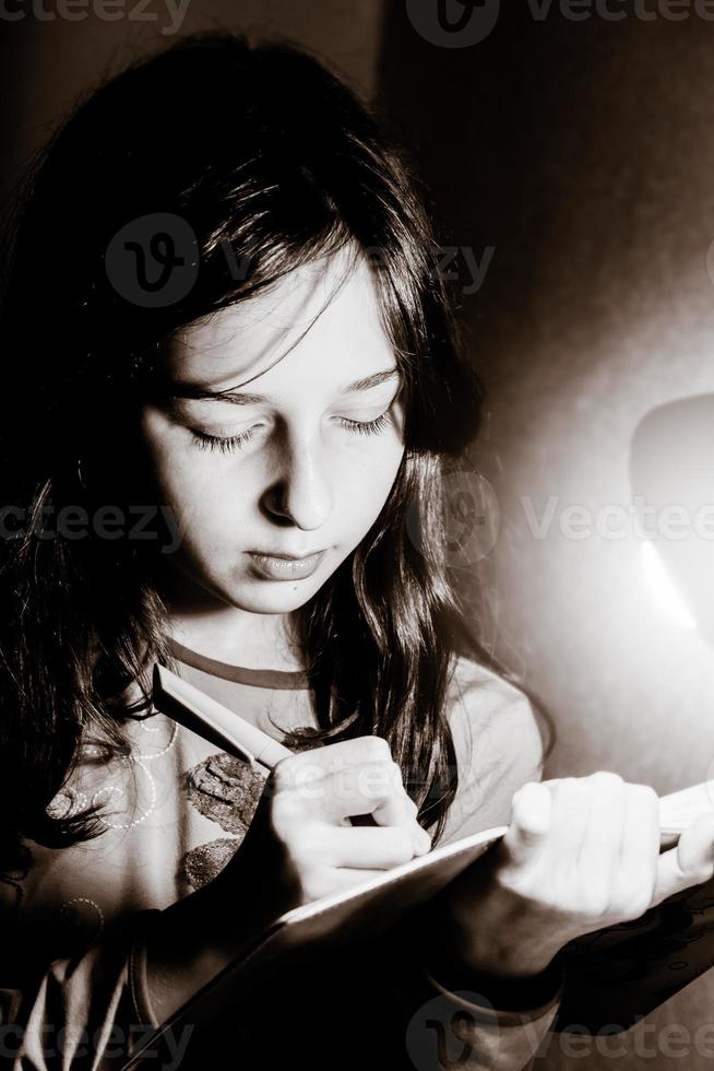 una adolescente escribe en un cuaderno con un bolígrafo. foto en blanco y negro de una niña iluminada por una lámpara de mesa.