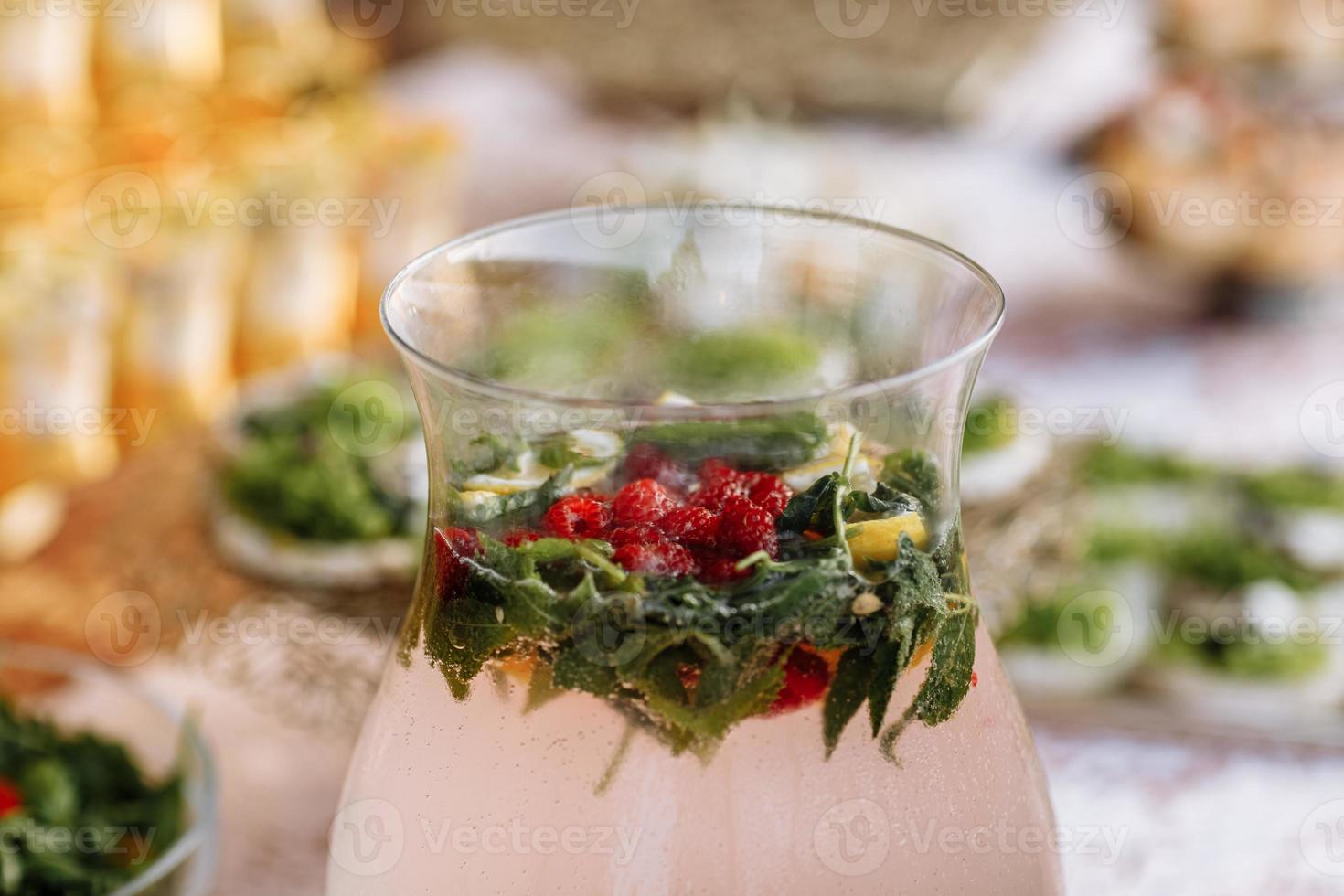 una gran jarra de vidrio llena de limonada se encuentra sobre una mesa de luz. en una jarra rodajas de fruta, naranja, limón, pomelo, frambuesa, menta. celebrando la fiesta foto