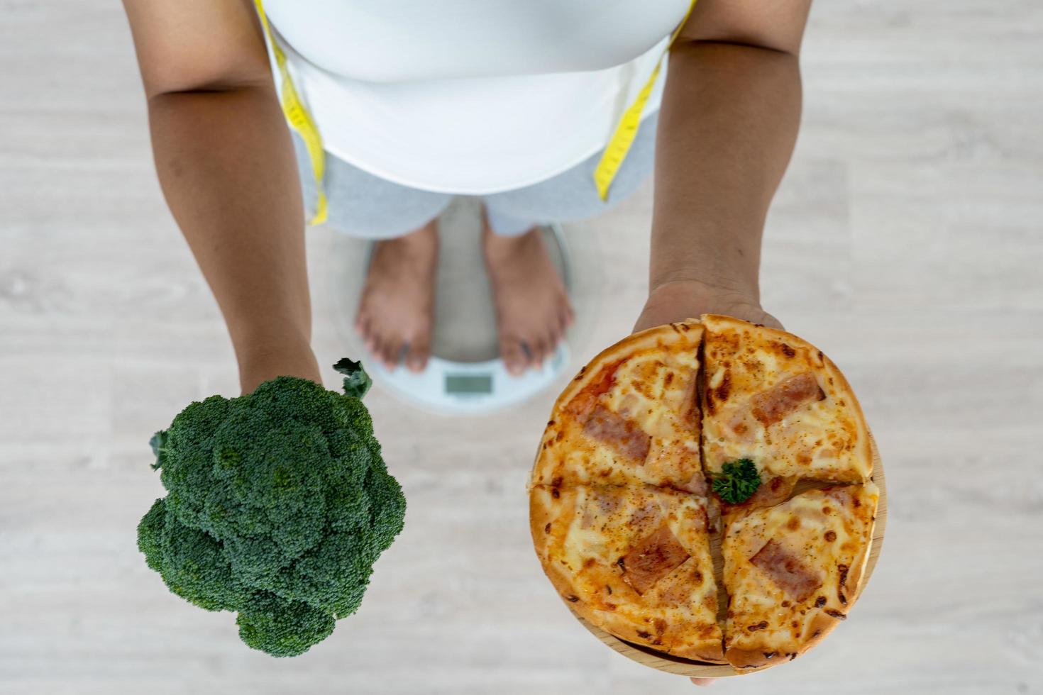 las mujeres se paran pesando en la balanza. la mano de una mujer sana se sostiene entre un brócoli y una pizza. decidir comer alimentos ricos en fibra y vitaminas para una buena salud foto