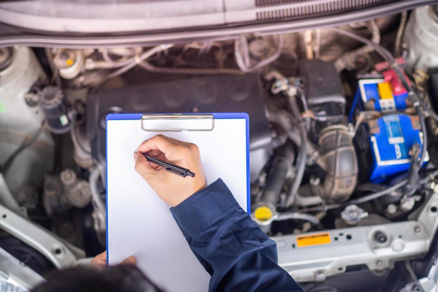 Hands of young professional mechanic in uniform writing on clipboard against car in open hood at the repair garage. Maintenance se. photo