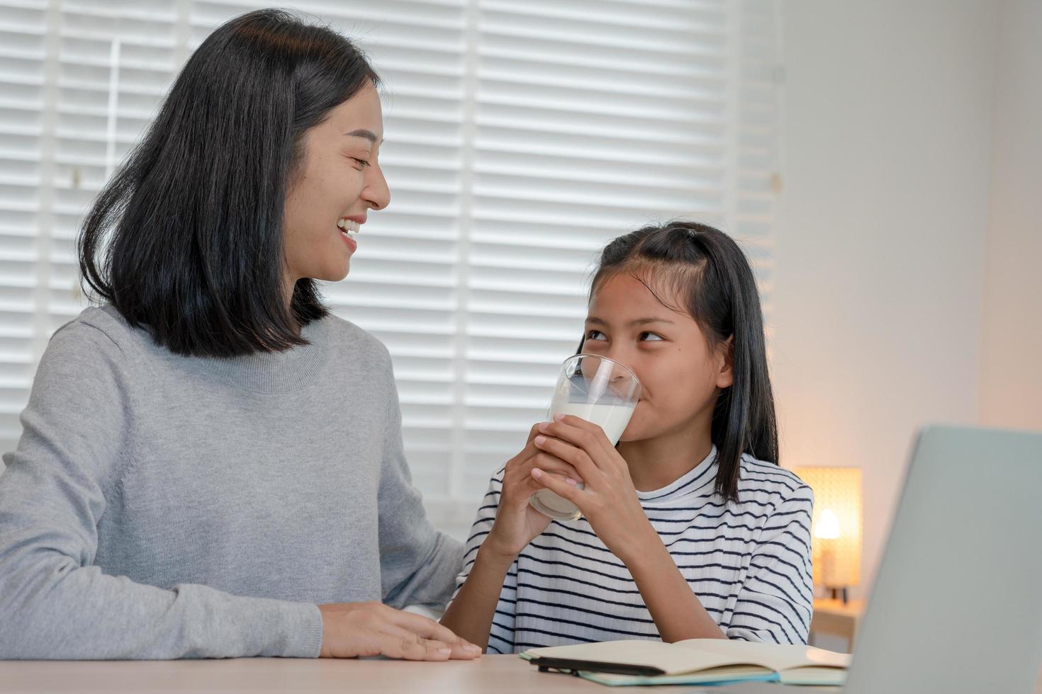 una niña asiática aprende en casa. hacer la tarea con la amable ayuda de la madre, animar para el examen. mamá le pasa un vaso de leche a su hija. niña feliz educación en el hogar. mamá enseña y aconseja educación juntos. foto