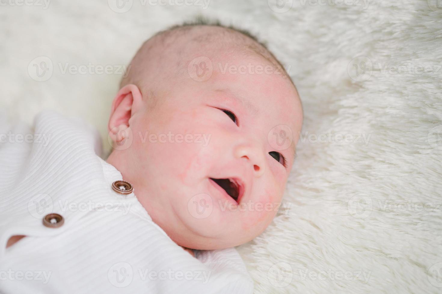 Closeup cute newborn baby in white bodysuit lying down alone on bed. Adorable infant rests on white bedsheets, staring at camera looking peaceful. Infancy, healthcare and paediatrics, babyhood concept photo