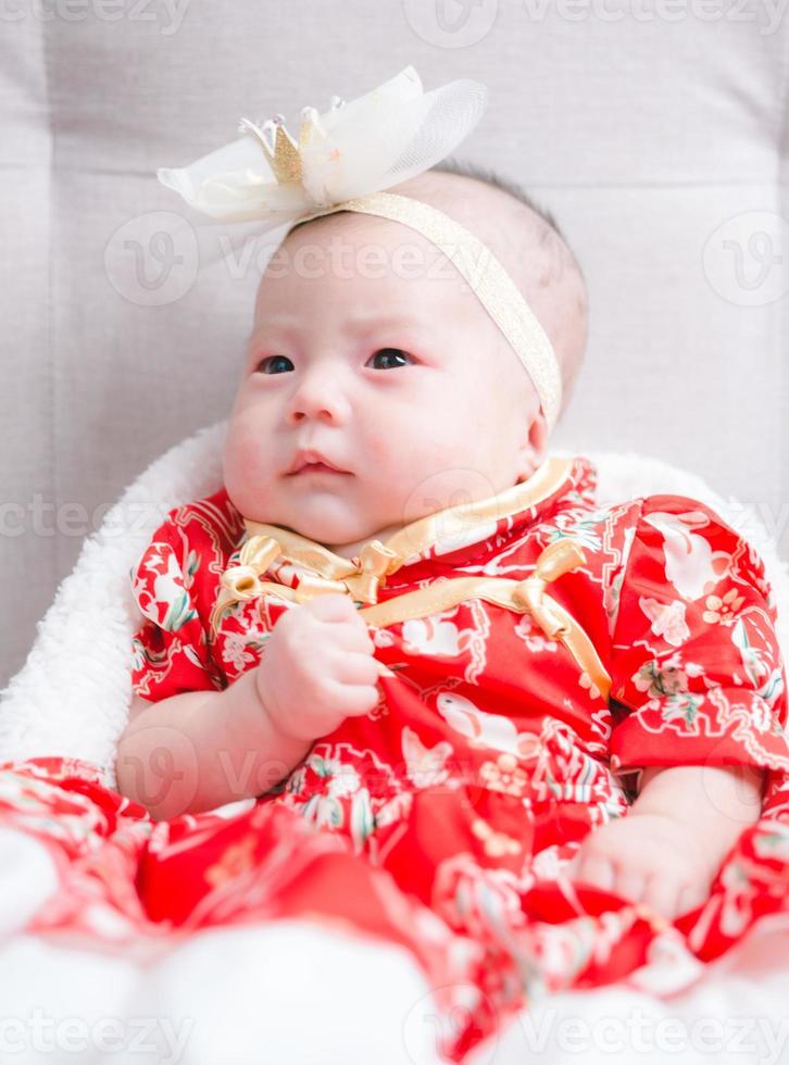 Closeup cute newborn baby in red bodysuit lying down alone on bed. Adorable infant rests on white bedsheets, staring at camera looking peaceful. Infancy, healthcare and paediatrics, babyhood concept. photo