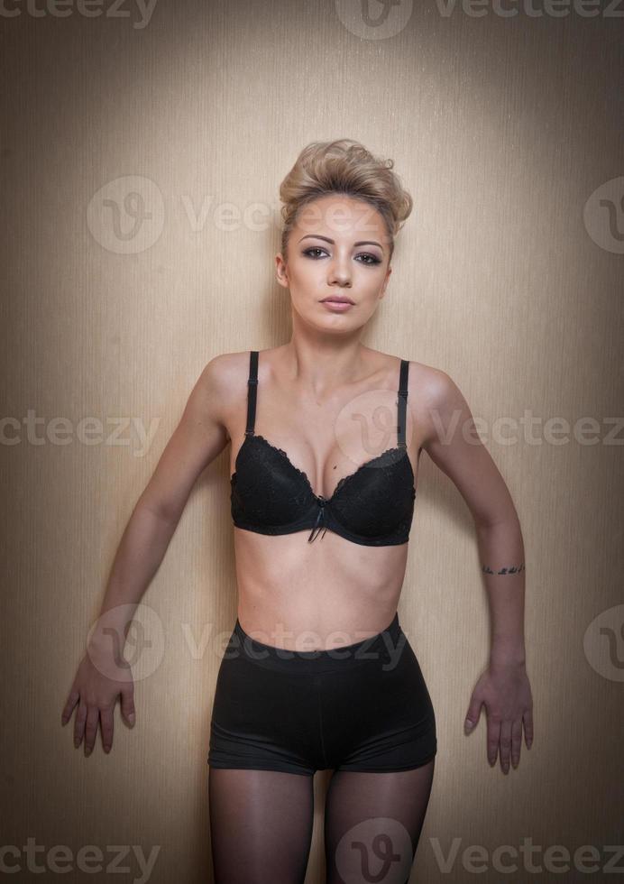 Attractive fair hair model with pantyhose and black bra posing provocatively. Fashion portrait of sensual short hair blonde, studio shot. Sensual female in black lingerie posing against wall. photo