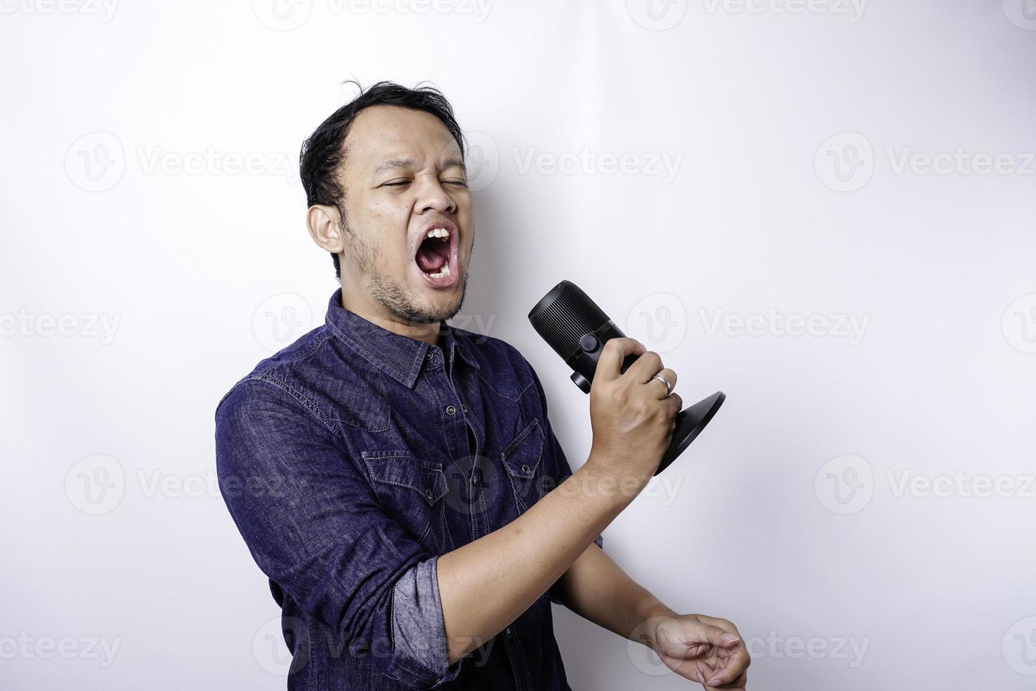 Portrait of carefree Asian man, having fun karaoke, singing in microphone while standing over white background photo