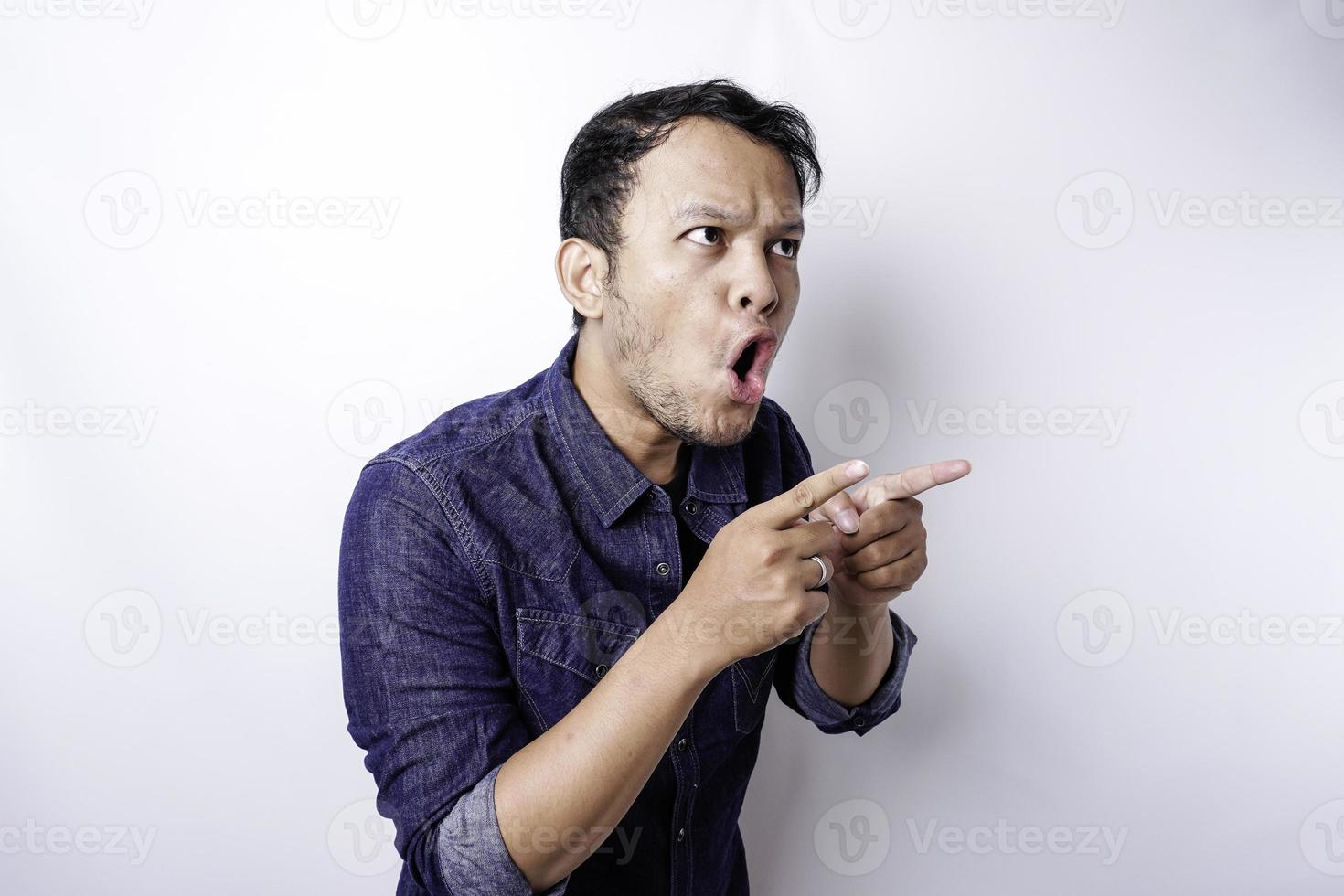 Shocked Asian man wearing blue shirt pointing at the copy space beside him, isolated by white background photo