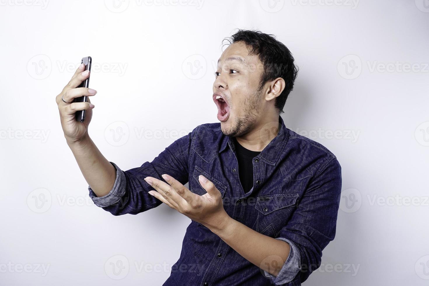 Shocked Asian man wearing blue shirt and holding his phone, isolated by white background photo