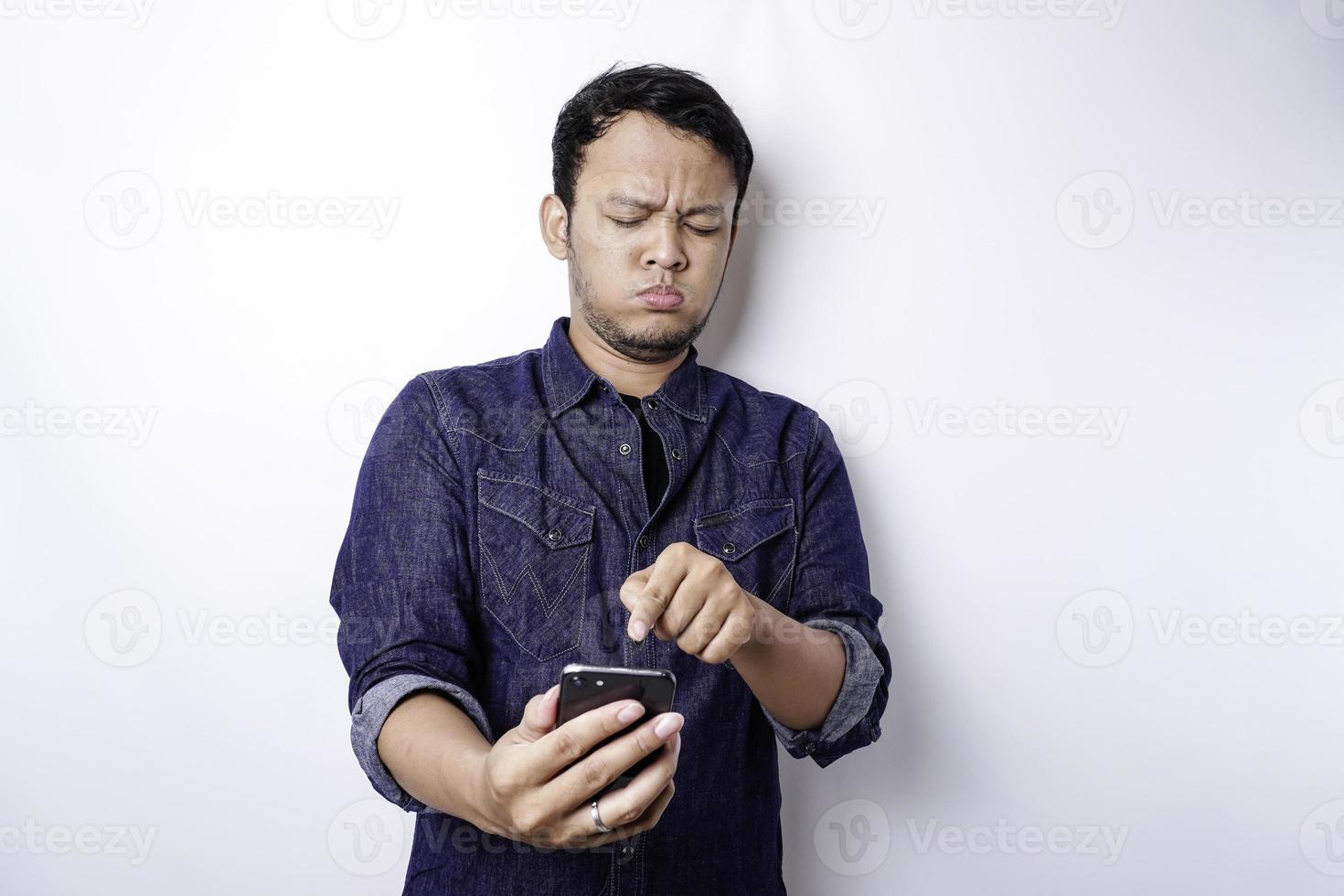 A thoughtful young Asian man is wearing blue shirt holding his phone and looks confused, isolated by white background photo