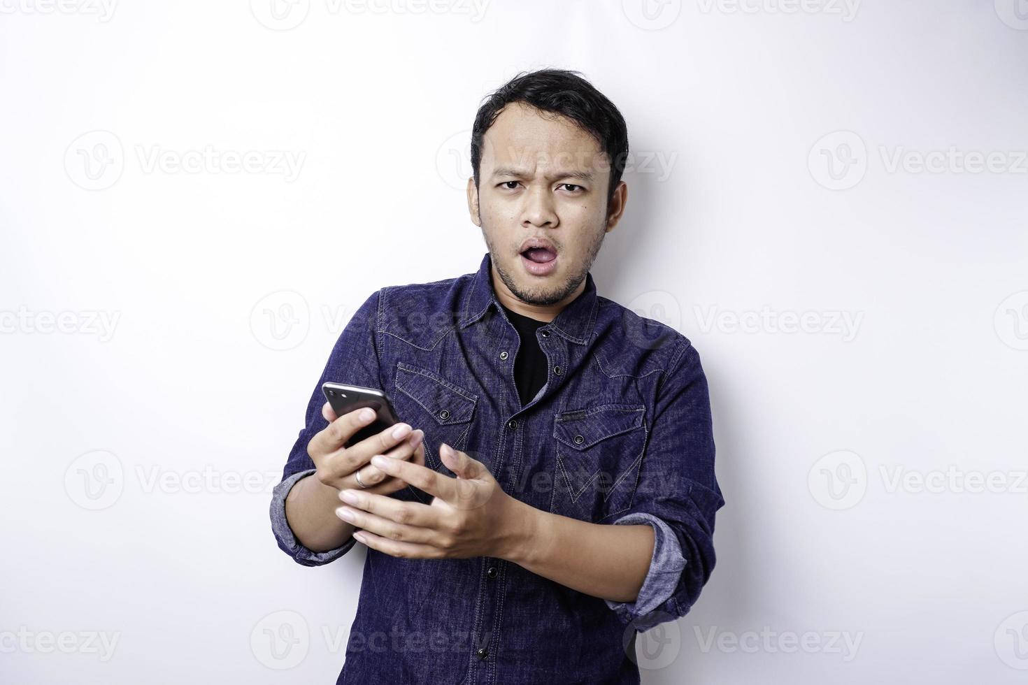 A thoughtful young Asian man is wearing blue shirt holding his phone and looks confused, isolated by white background photo