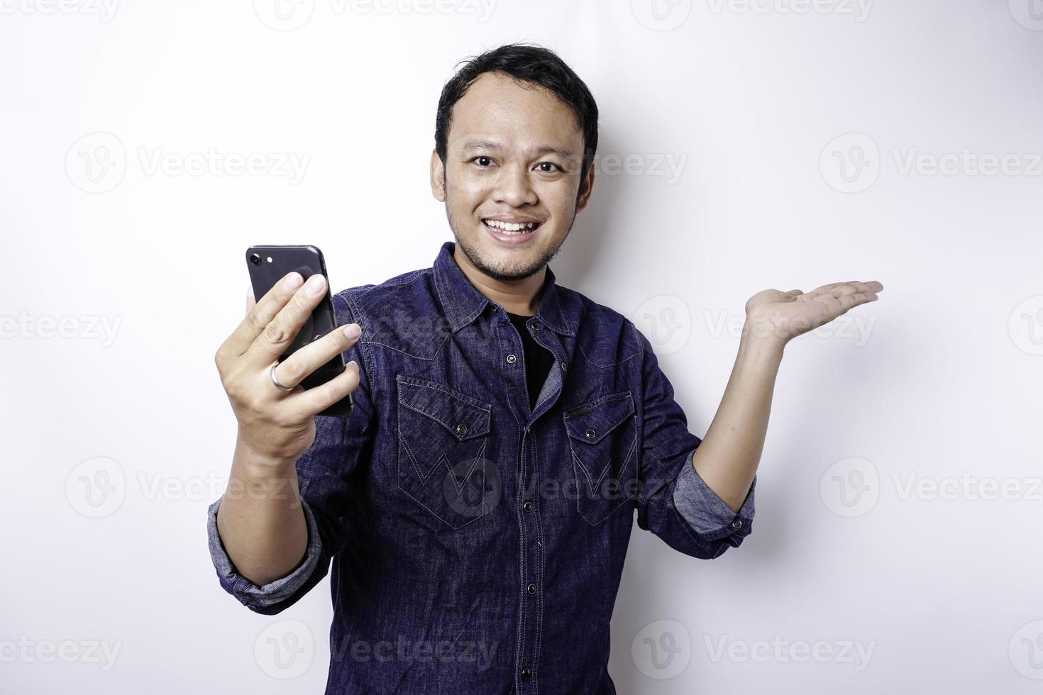 Excited Asian man wearing blue shirt pointing at the copy space beside him while holding his phone, isolated by white background photo