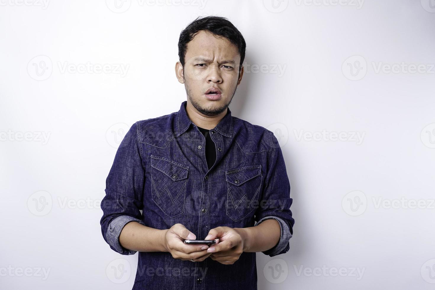 A thoughtful young Asian man is wearing blue shirt holding his phone and looks confused, isolated by white background photo