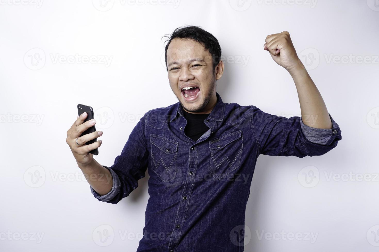 A young Asian man with a happy successful expression wearing blue shirt and holding his phone, isolated by white background photo