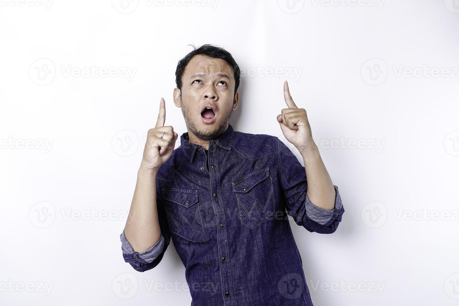 Shocked Asian man wearing blue shirt pointing at the copy space on top of him, isolated by white background photo