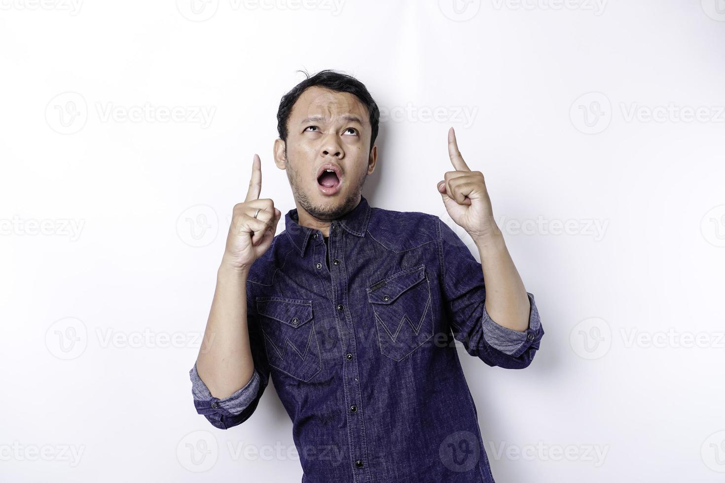 hombre asiático conmocionado con camisa azul apuntando al espacio de copia encima de él, aislado por fondo blanco foto