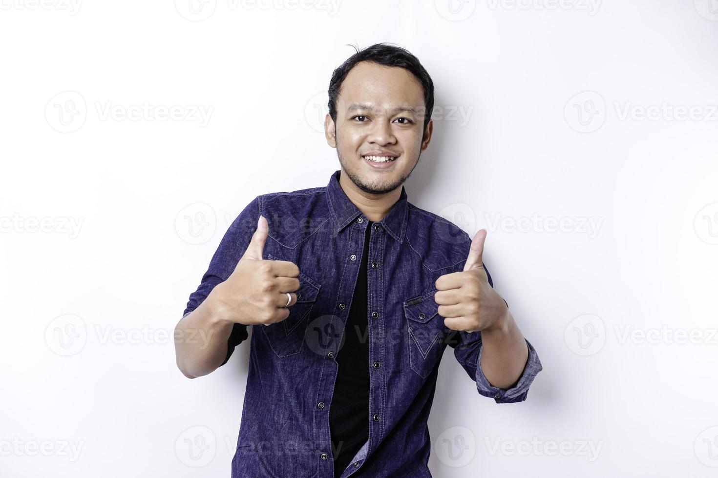 Excited Asian man wearing blue shirt gives thumbs up hand gesture of approval, isolated by white background photo