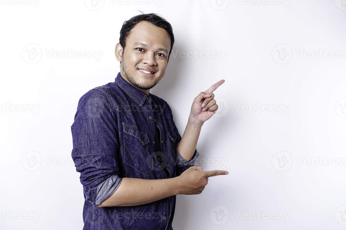 Excited Asian man wearing blue shirt pointing at the copy space beside him, isolated by white background photo