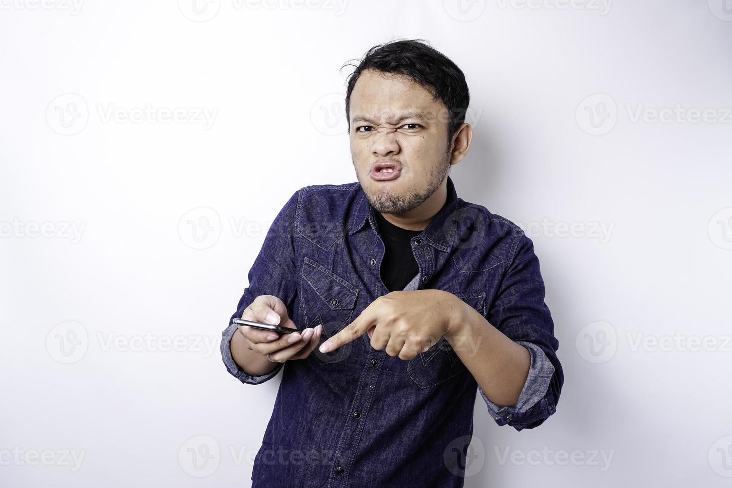 The angry and mad face of Asian man in blue shirt holding his phone isolated white background. photo