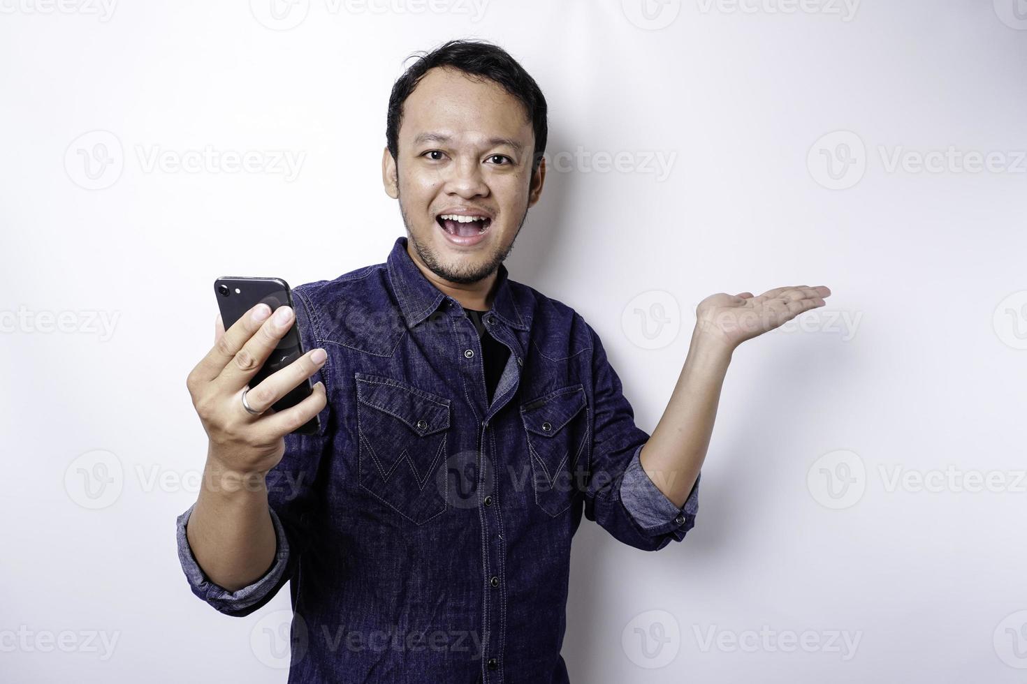 Excited Asian man wearing blue shirt pointing at the copy space beside him while holding his phone, isolated by white background photo