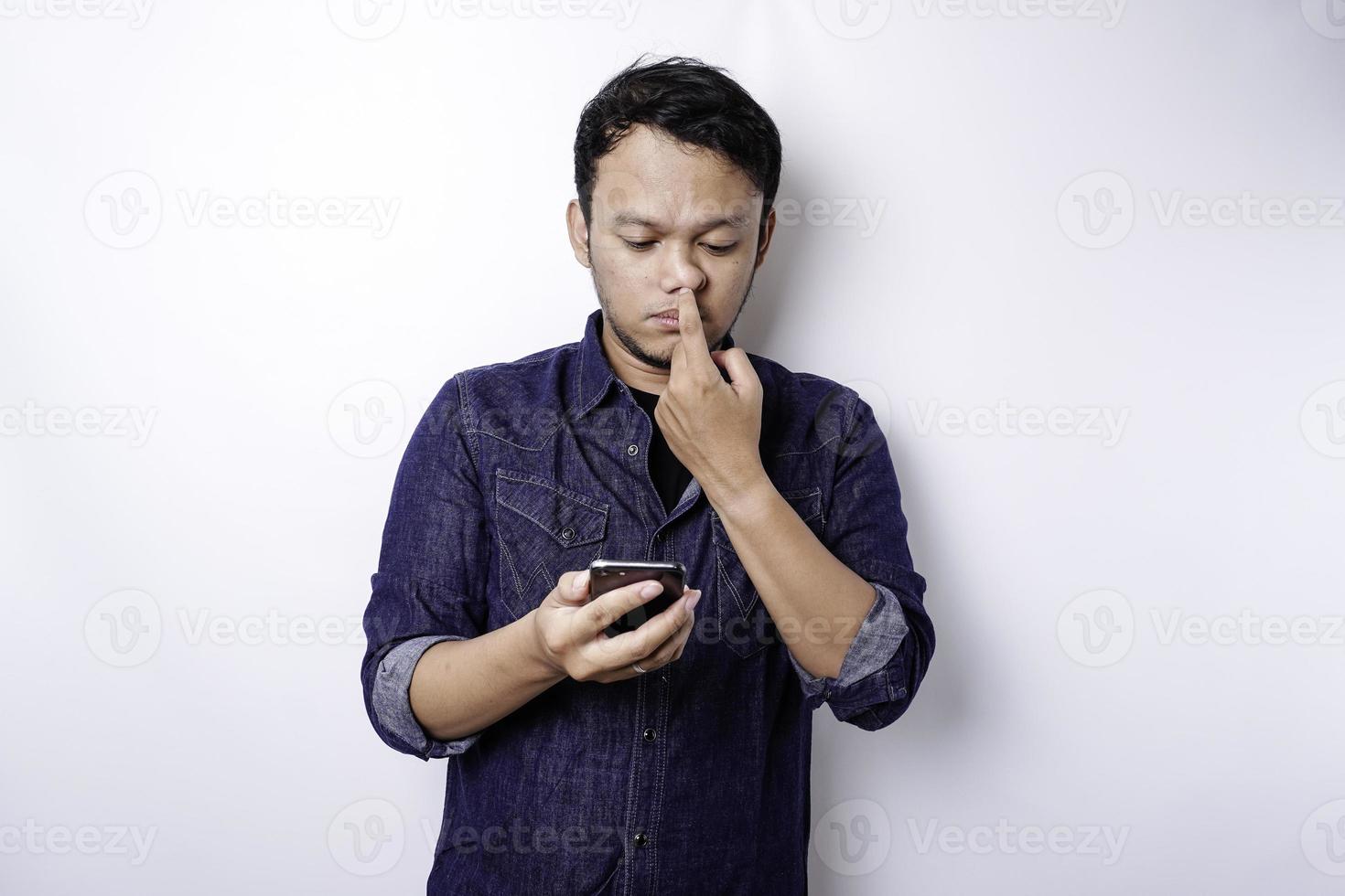 A thoughtful young man dressed in blue shirt while looking aside holding his phone, isolated by white background photo